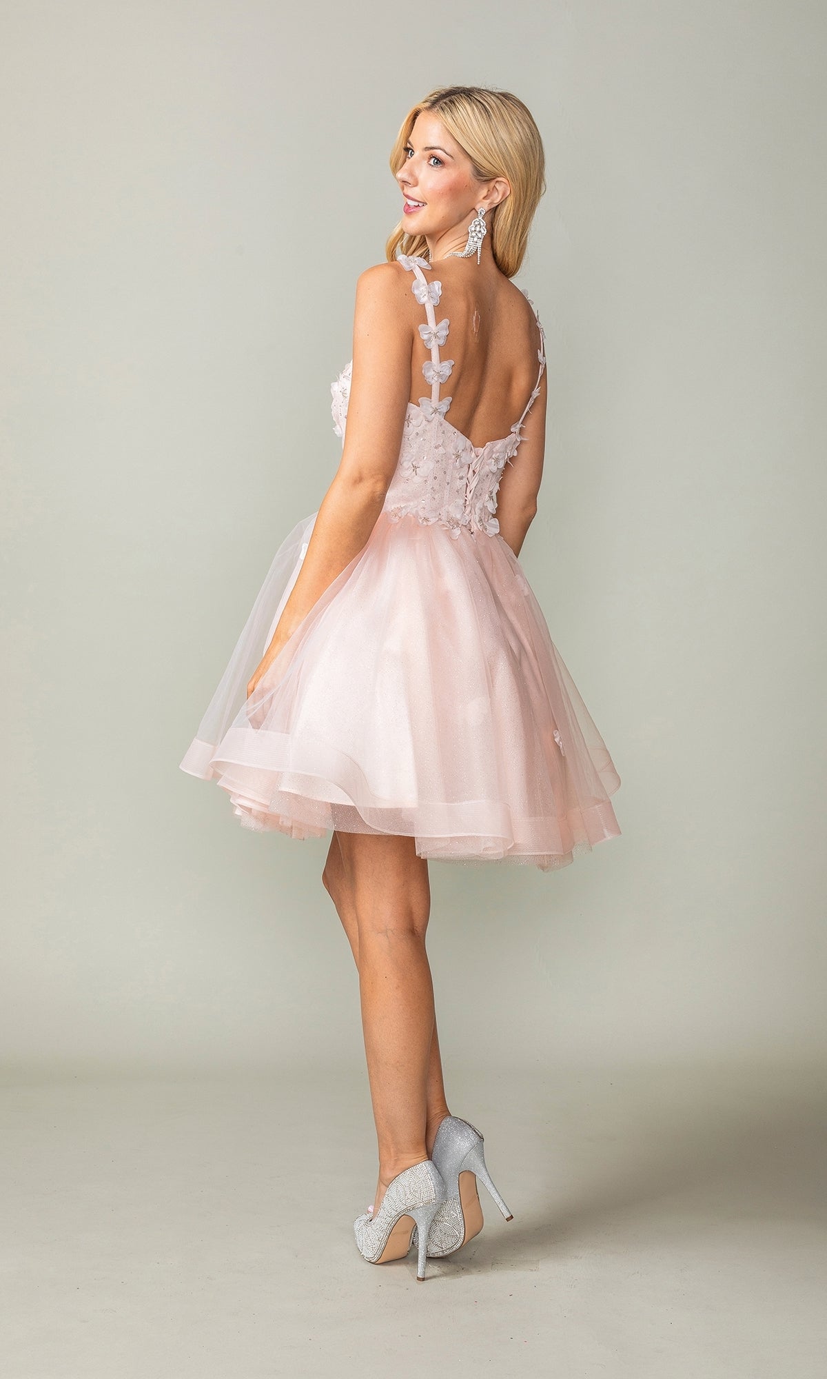 Woman in a Lace-Up Short Butterfly Homecoming Dress 3369, featuring pale pink layered tulle, 3-D butterflies, and a sleeveless design, wears silver heels and stands looking over her shoulder.